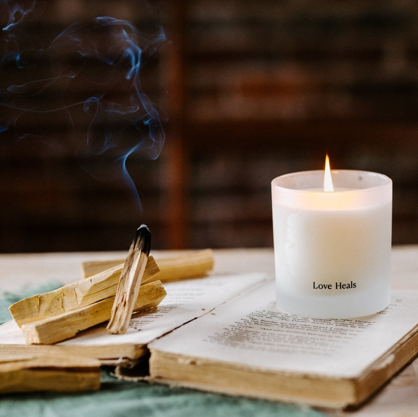 A lit frosted Palo Santo candle sits on top of an open antique book with worn paper edges. The other page carries the Palo Santo wood that has a charred piece emitting smoke.