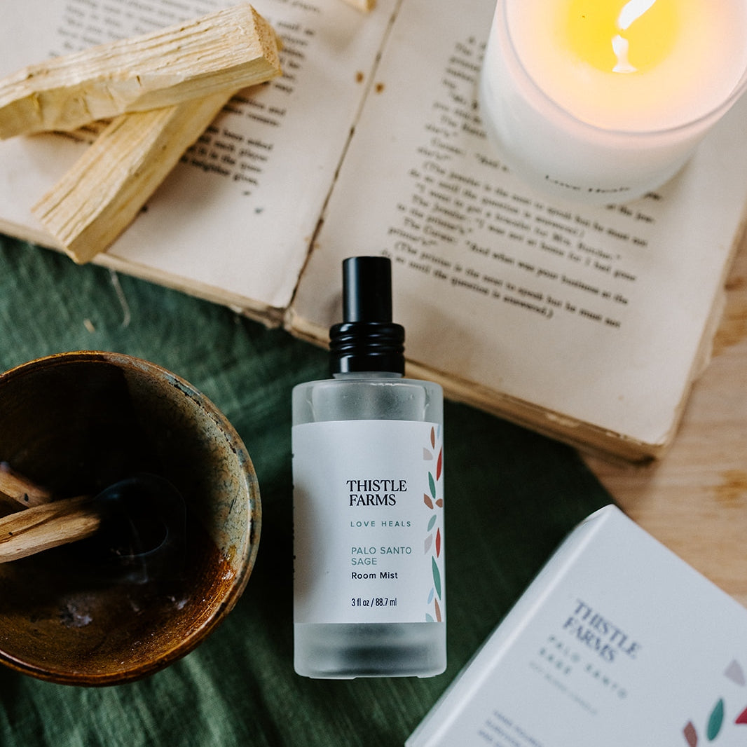 A Frosted spray bottle of Palo Santo Sage Room Mist lays on a rich green textile surrounded by a lit candle, palo santo wood, and an antique book with worn pages.