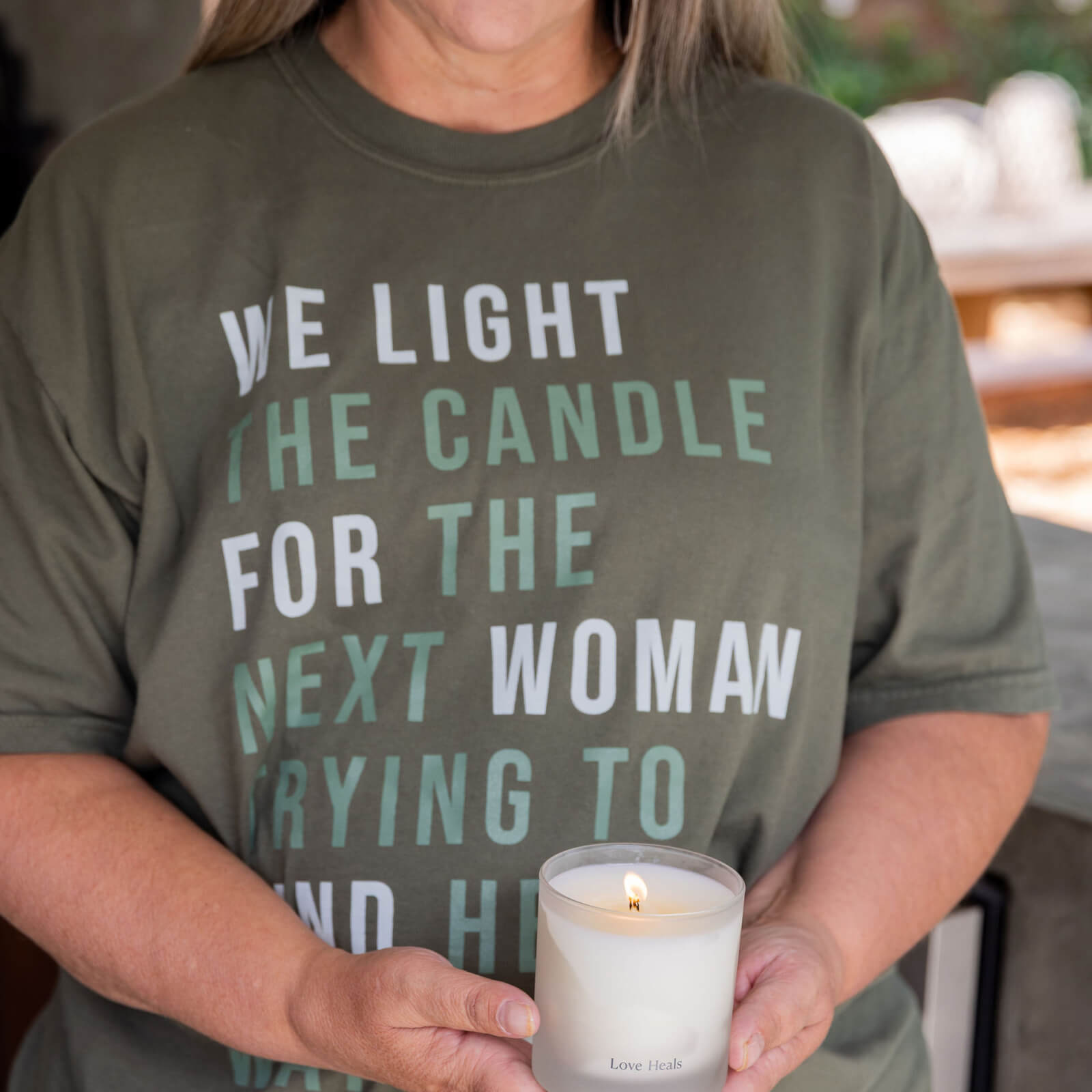 Person wearing a green t-shirt with a message "WE LIGHT THE CANDLE FOR THE NEXT WOMAN TRYING TO FIND HER WAY HOME", holding a candle.