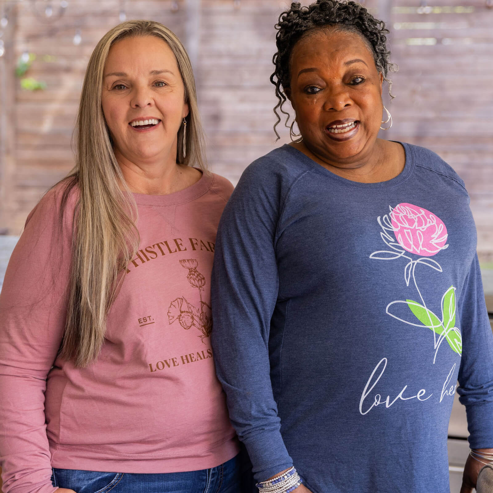 Two women wearing pink and blue Thistle Farms branded shirts with floral designs and text.