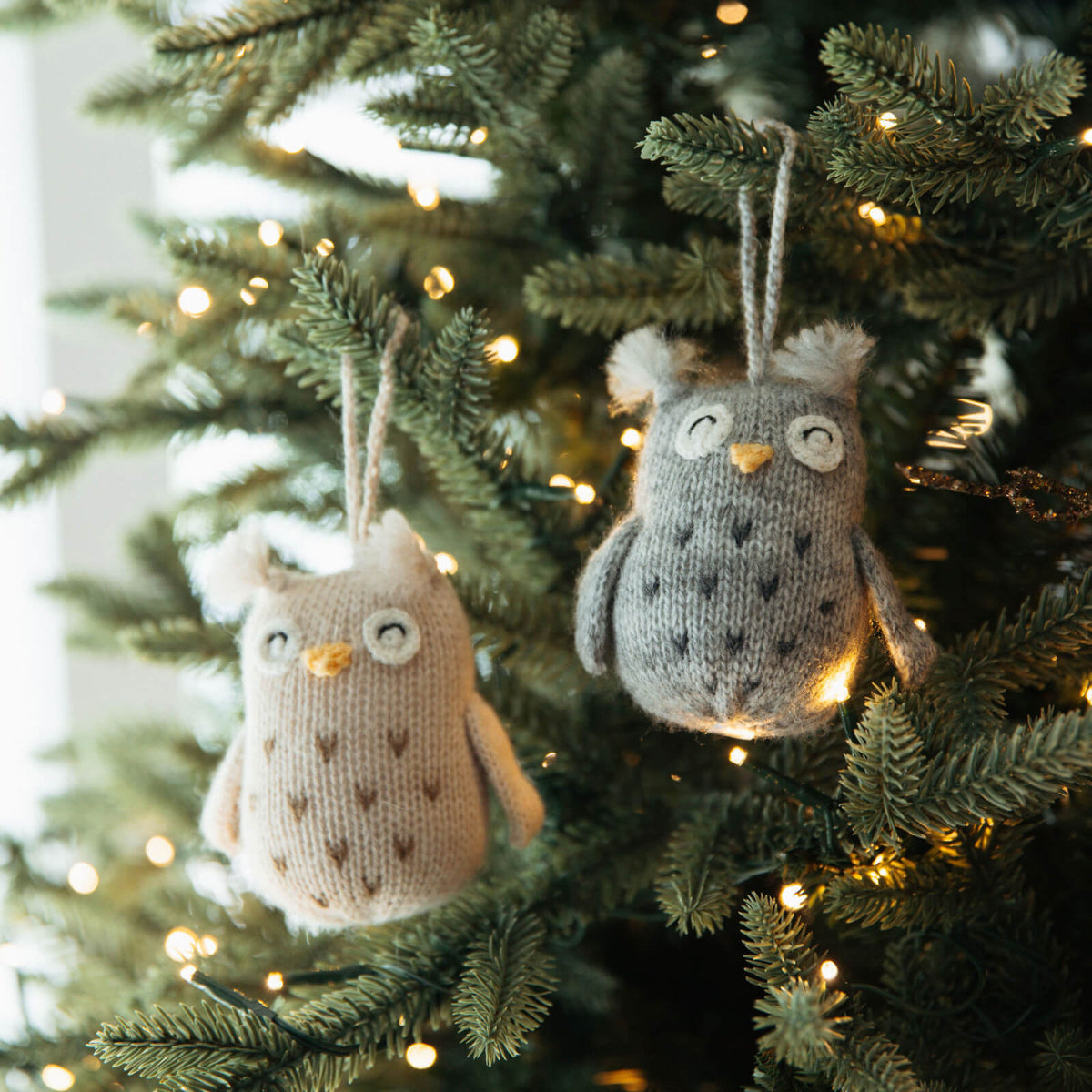 Two knitted owl ornaments hanging on a decorated Christmas tree.