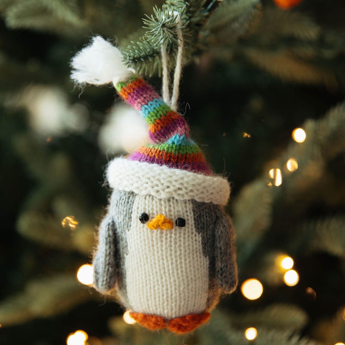 Knitted penguin ornament with a colorful hat hanging on a Christmas tree.