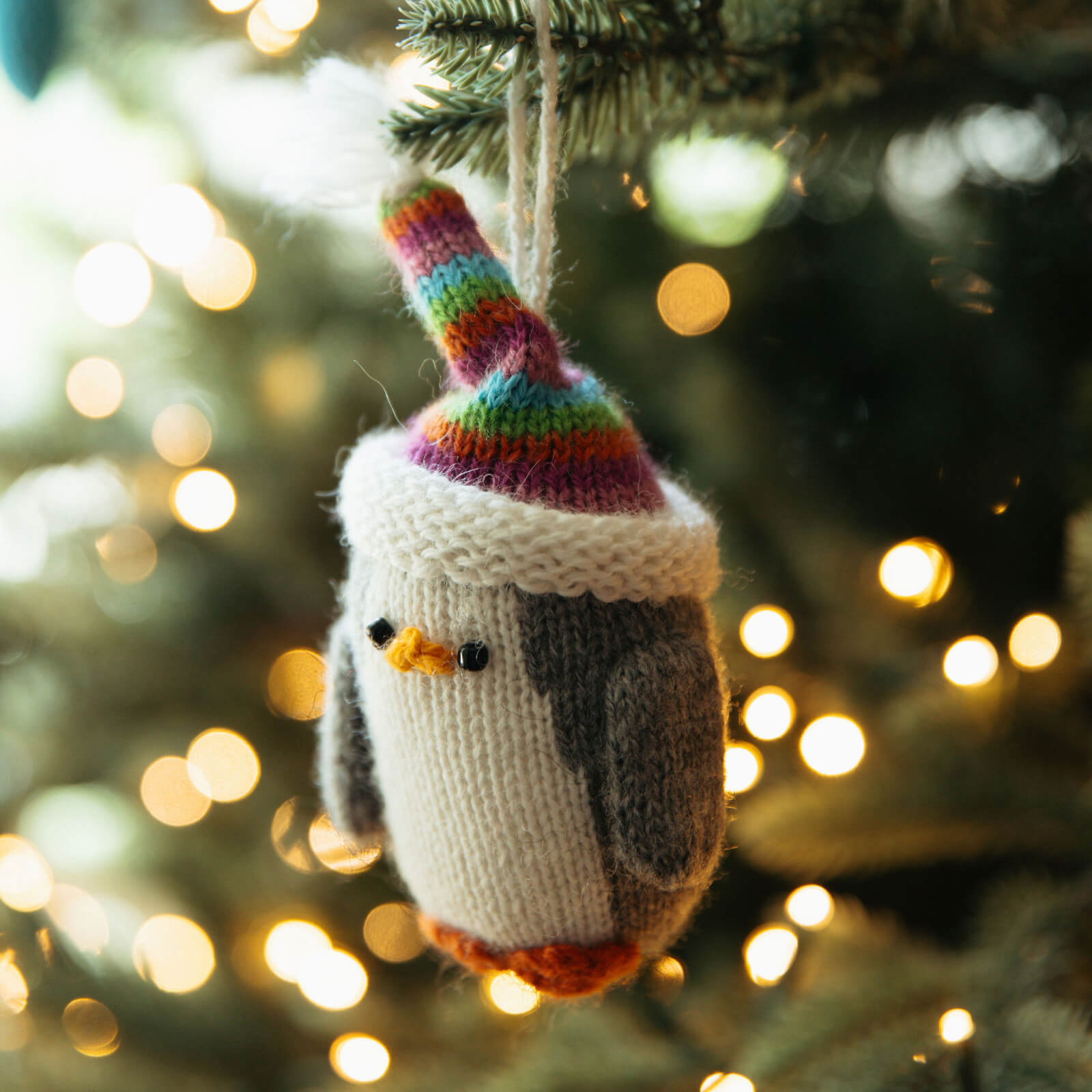 Knitted penguin ornament with a colorful hat hanging on a Christmas tree with blurred lights in the background.