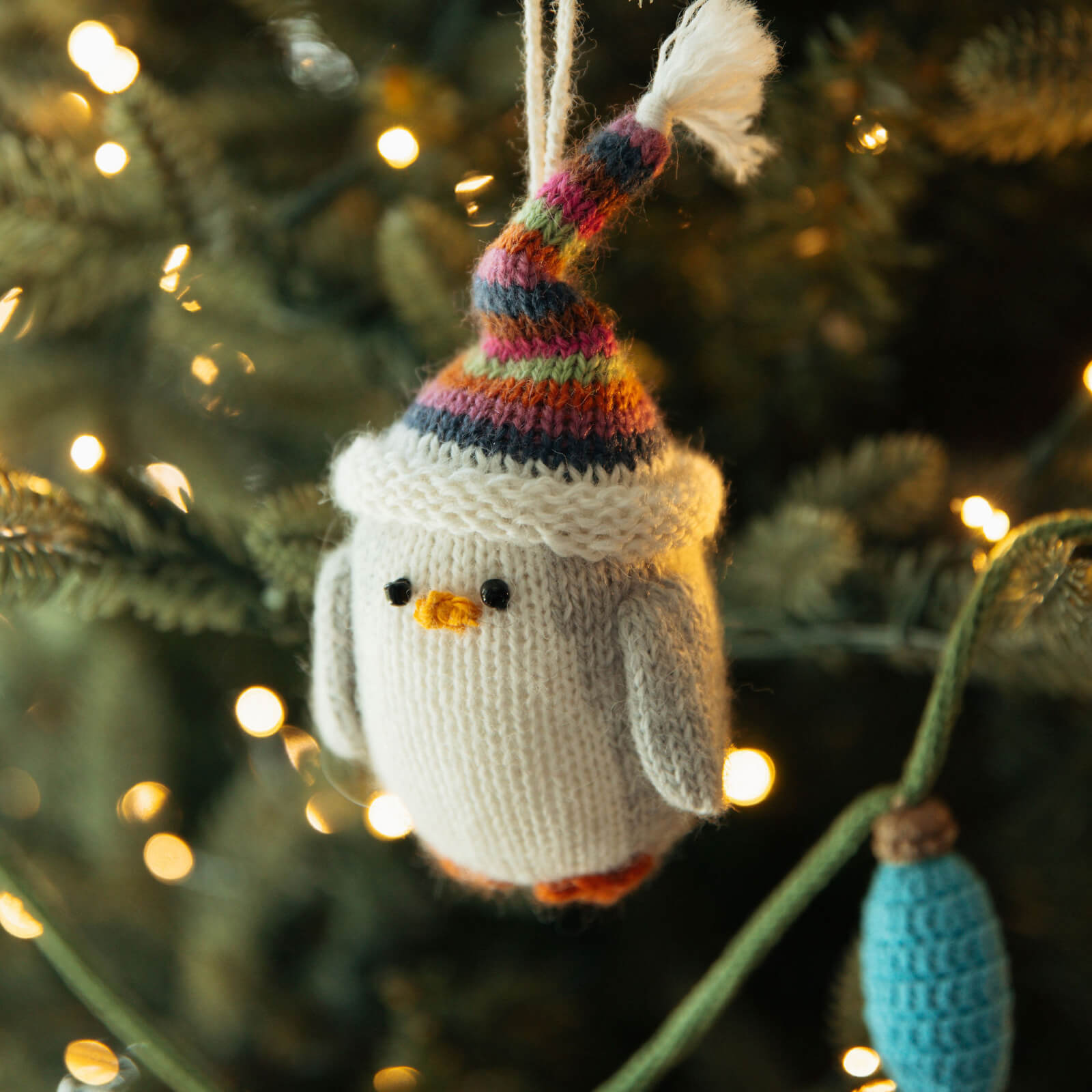 Knitted penguin ornament with a colorful hat hanging on a Christmas tree.