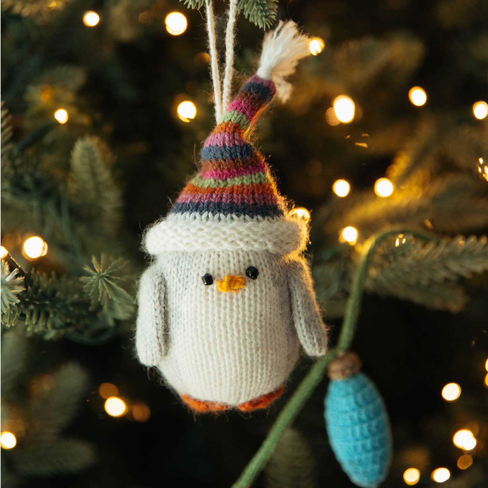 Knitted penguin ornament with a colorful hat hanging on a Christmas tree.