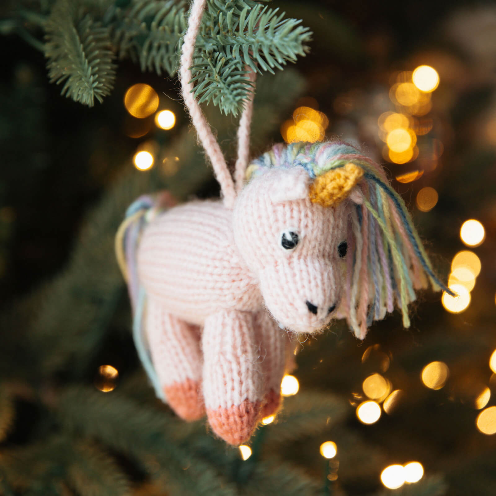 Knitted unicorn ornament hanging on a Christmas tree with blurred lights in the background