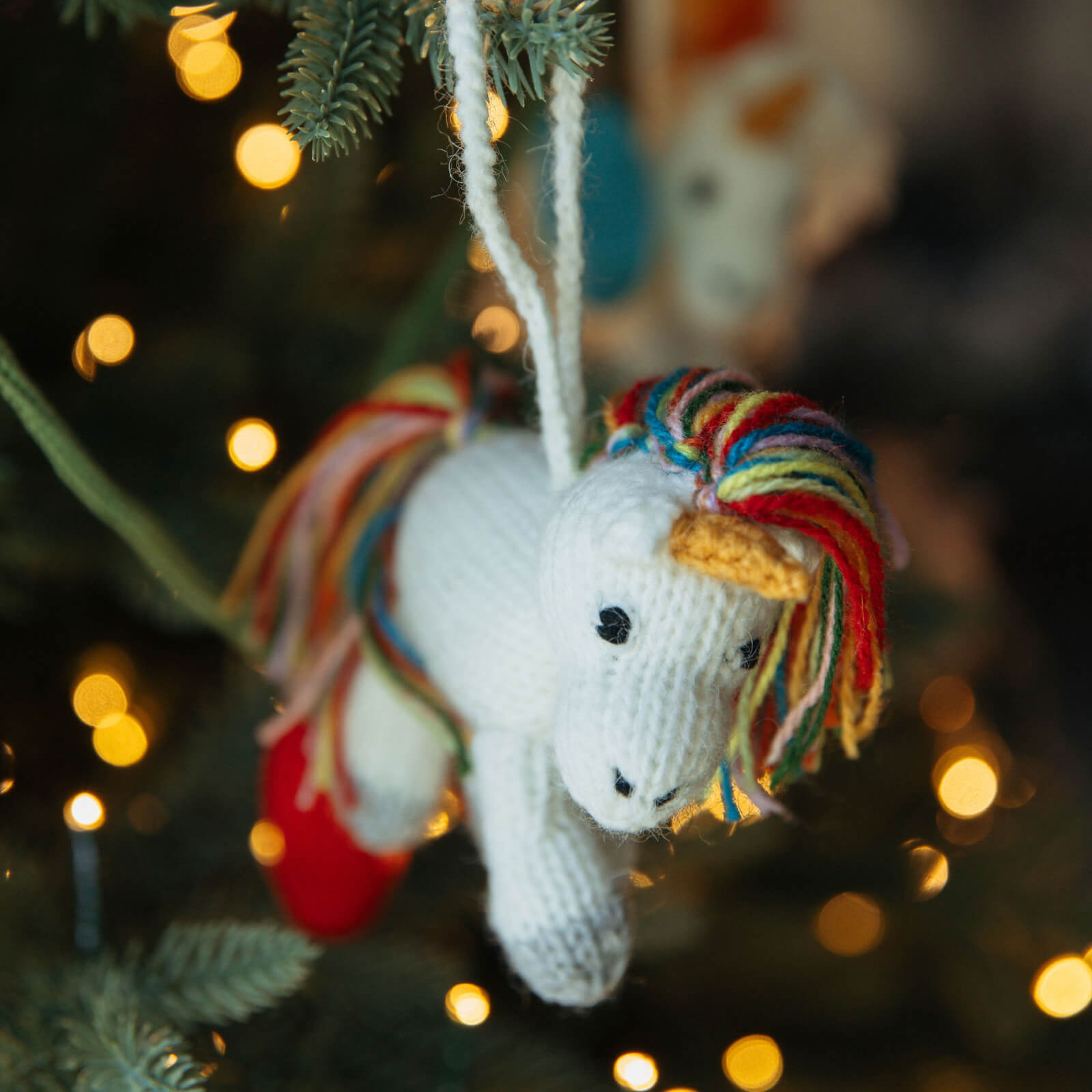 Knitted unicorn ornament with colorful mane hanging on a Christmas tree with blurred lights in the background.