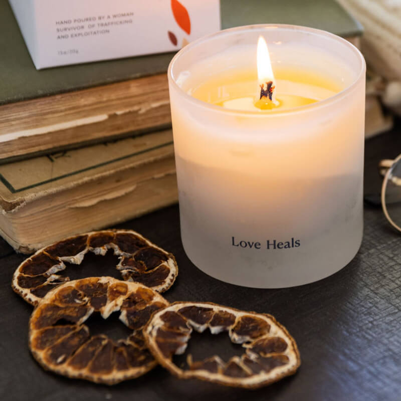 Frosted glass candle that is lit with bright flame. Dried oranges are laid against the candle on the dark wood right by some old books.