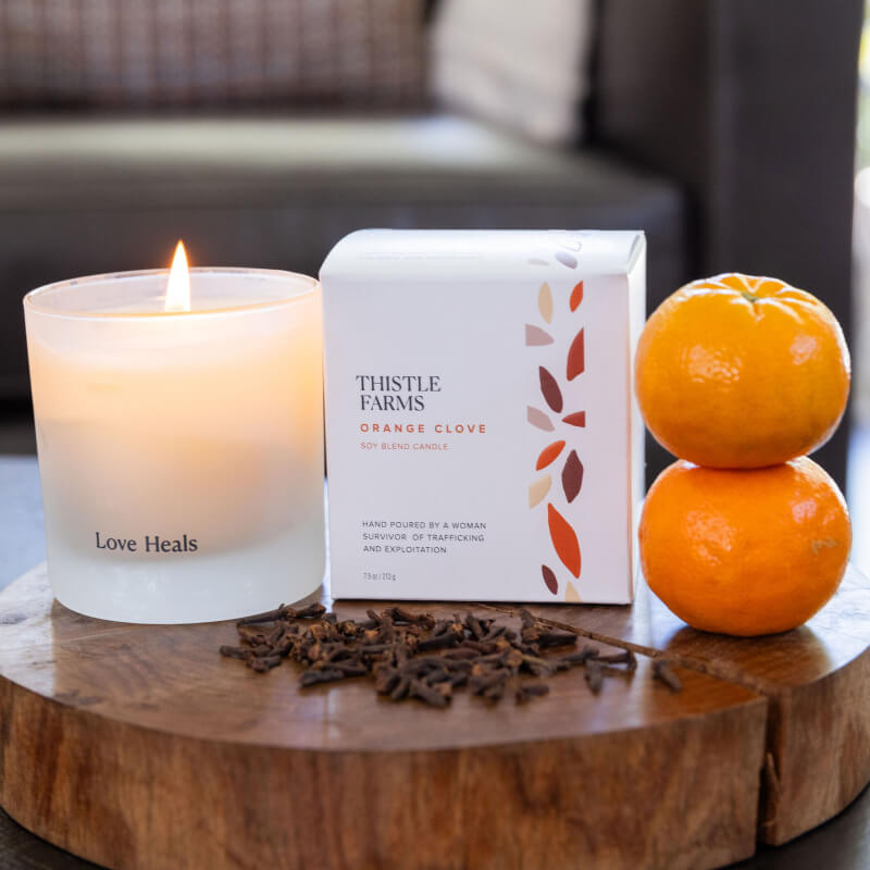 Lit Orange Clove candle alongside box and oranges stacked on top of each other. It is sitting on top of a circular wood cut piece.