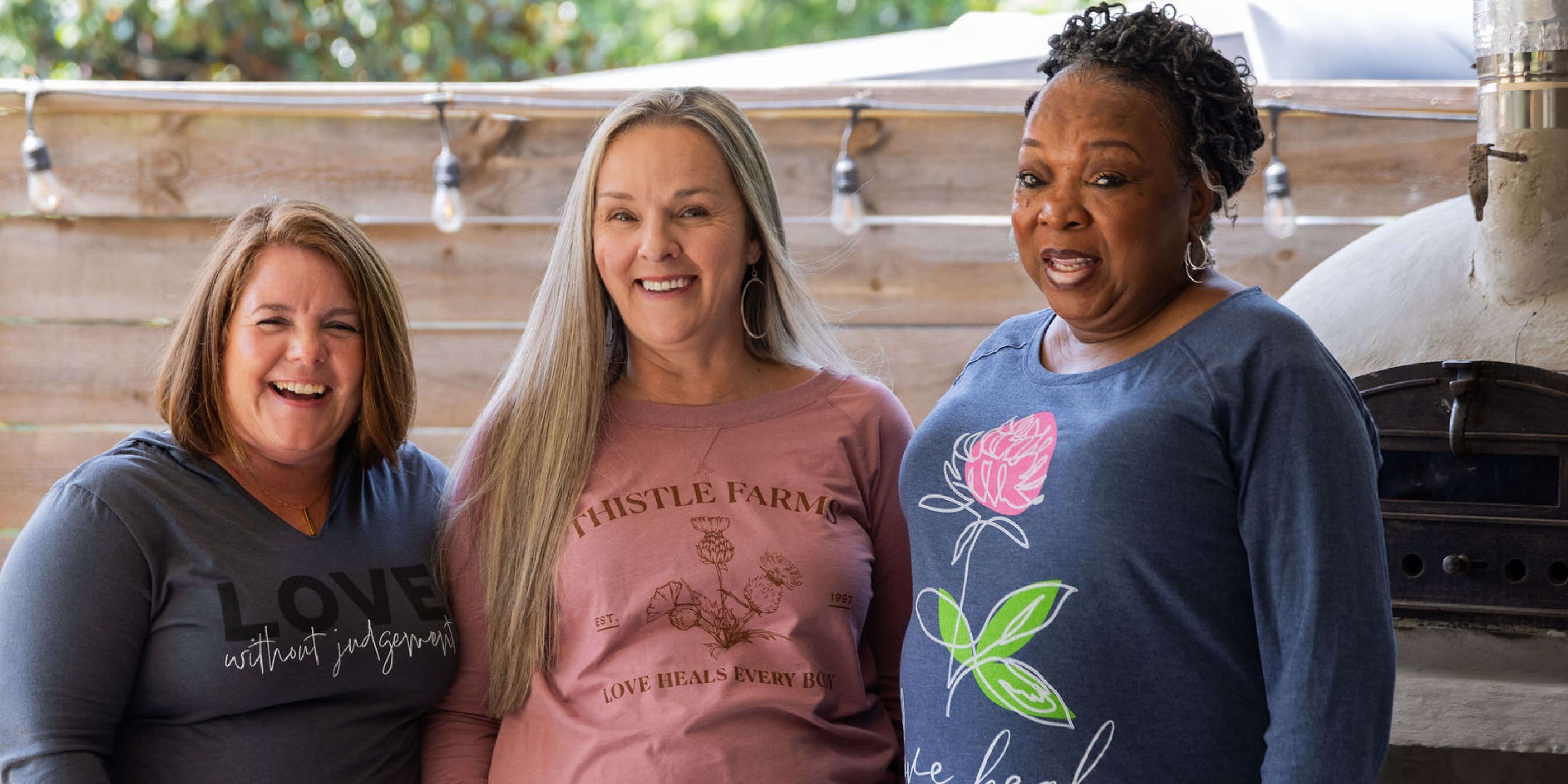 Three women standing together outdoors, wearing branded shirts.