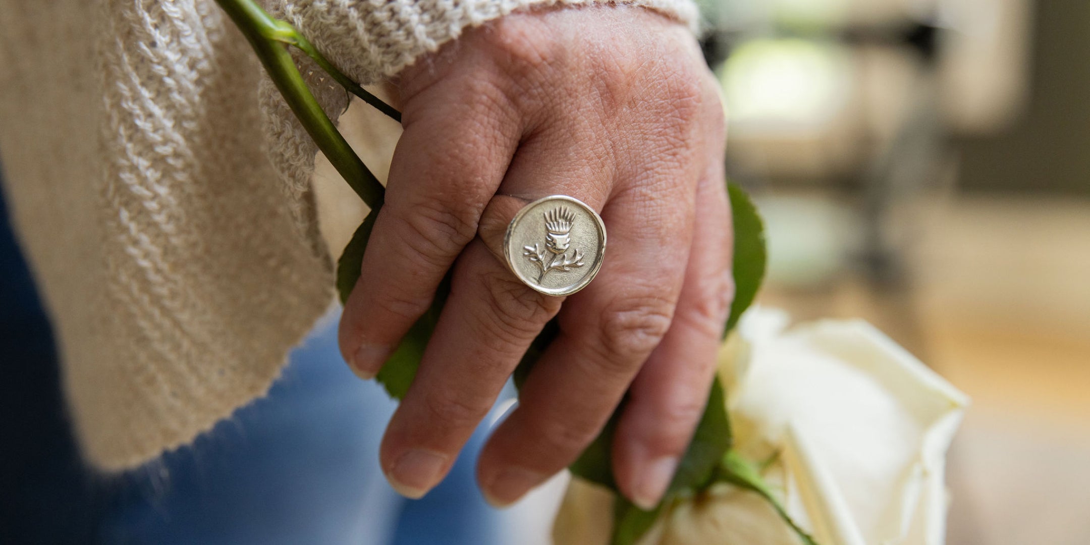 Hand holding a white rose with a Thistle Farms Signet Ring on a blurred background