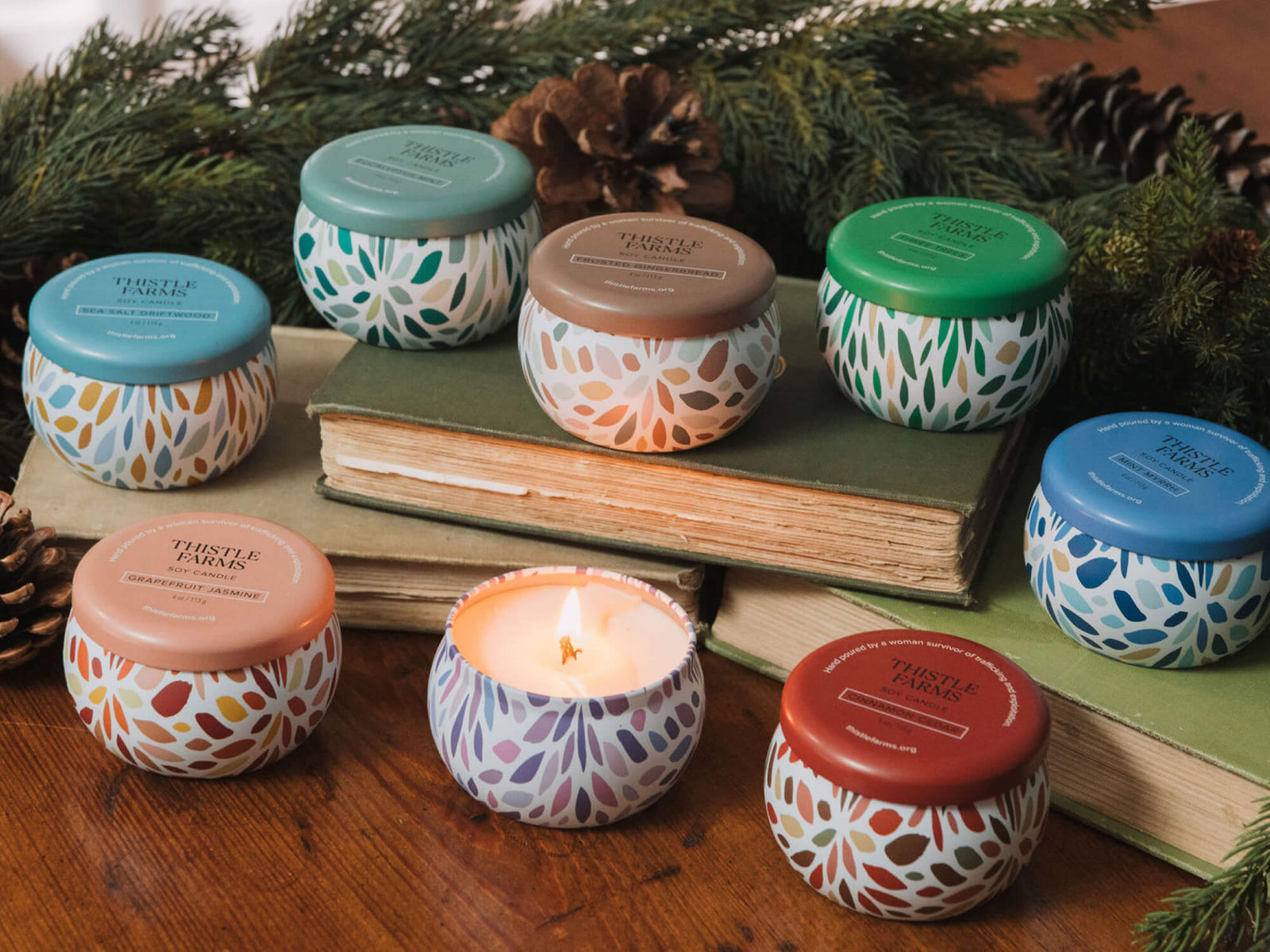 Assorted small candles with colorful lids on a wooden surface with books and pinecones in the background.