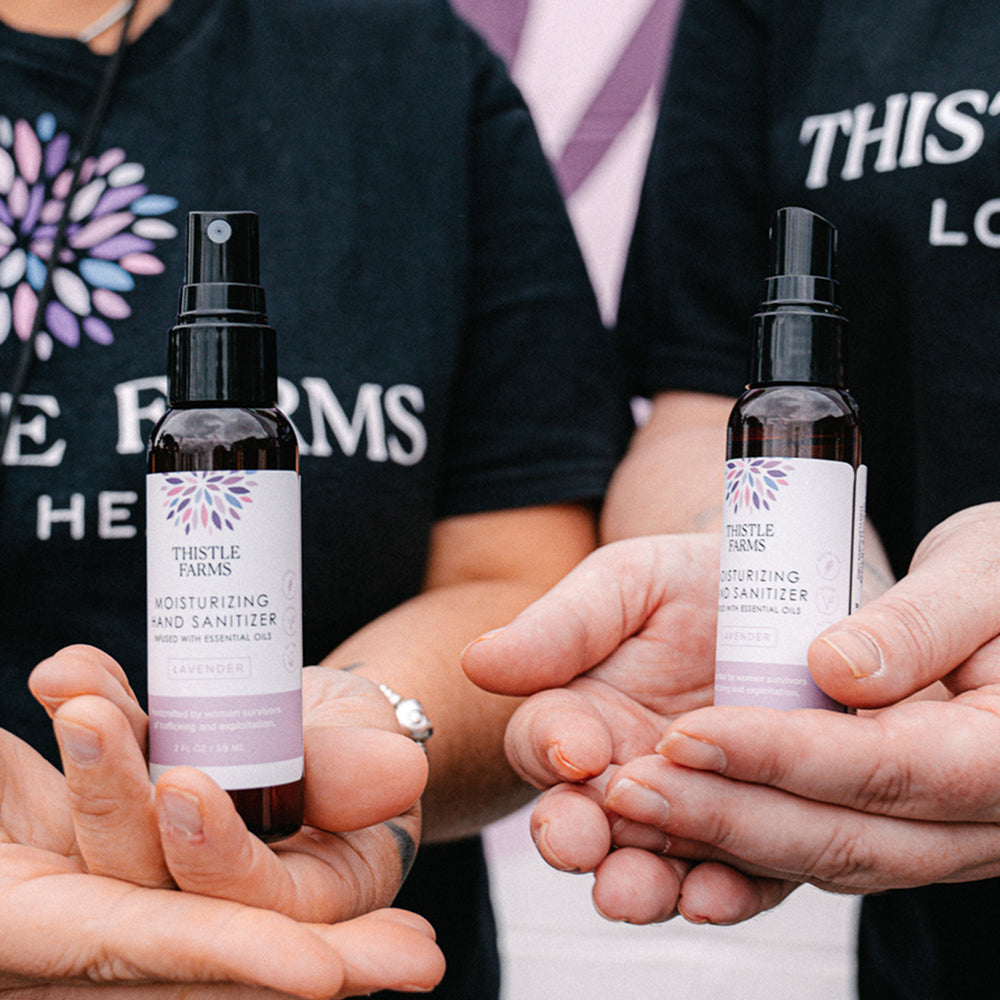 Two people holding bottles of Thistle Farms lavender hand sanitizer with a blurred background