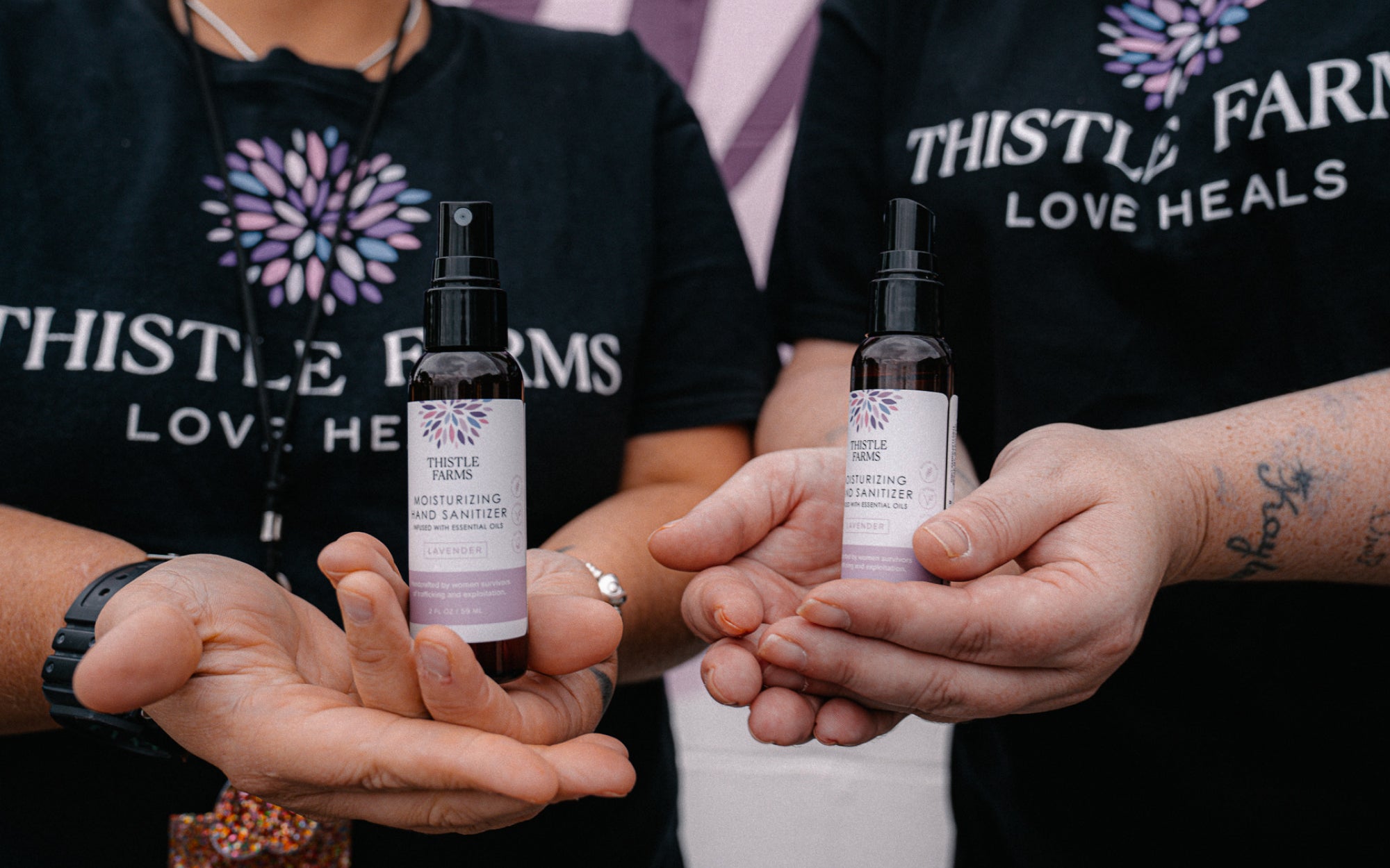 Two people holding bottles of Thistle Farm lavender hand sanitizer with visible branding.