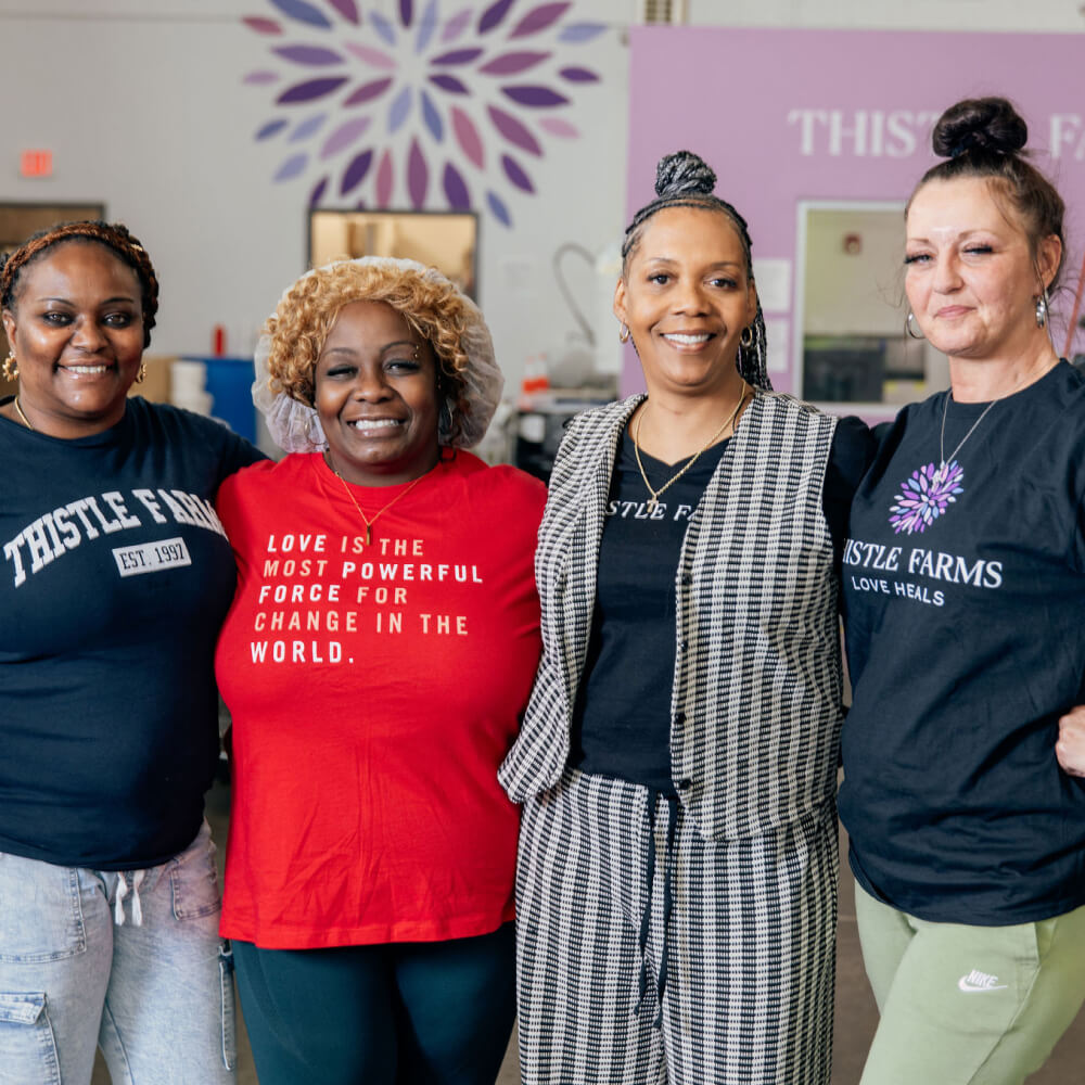 Four women survivors posing together, three wearing 'Thistle Farms' shirts, in a Thistle Farms Manufacuring Warehouse.