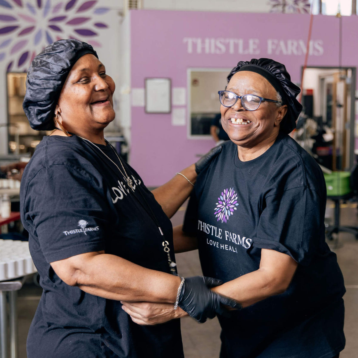 Two women wearing Thistle Farms shirts in a kitchen setting