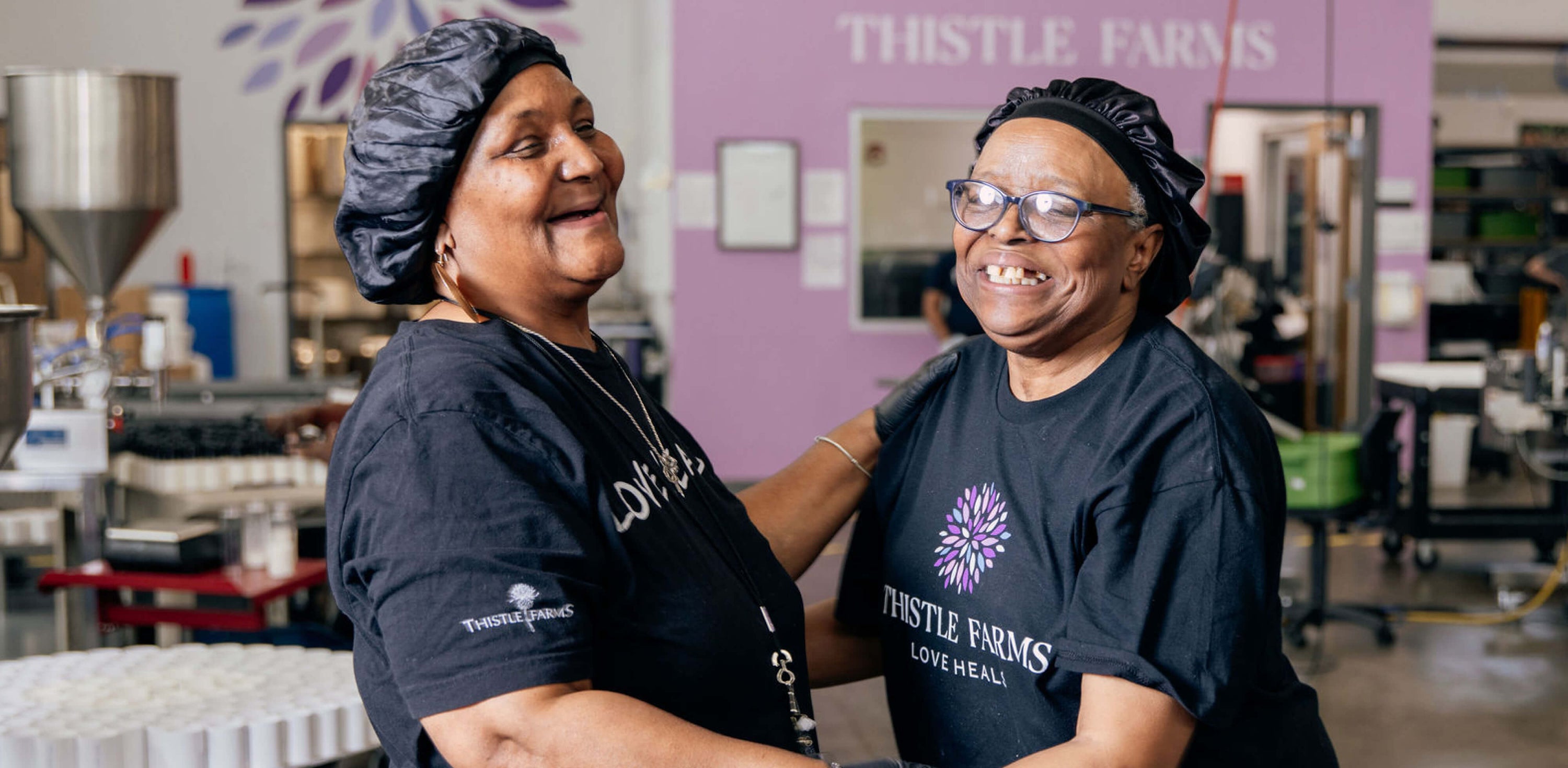 Two women wearing Thistle Farms t-shirts in a warehouse setting.