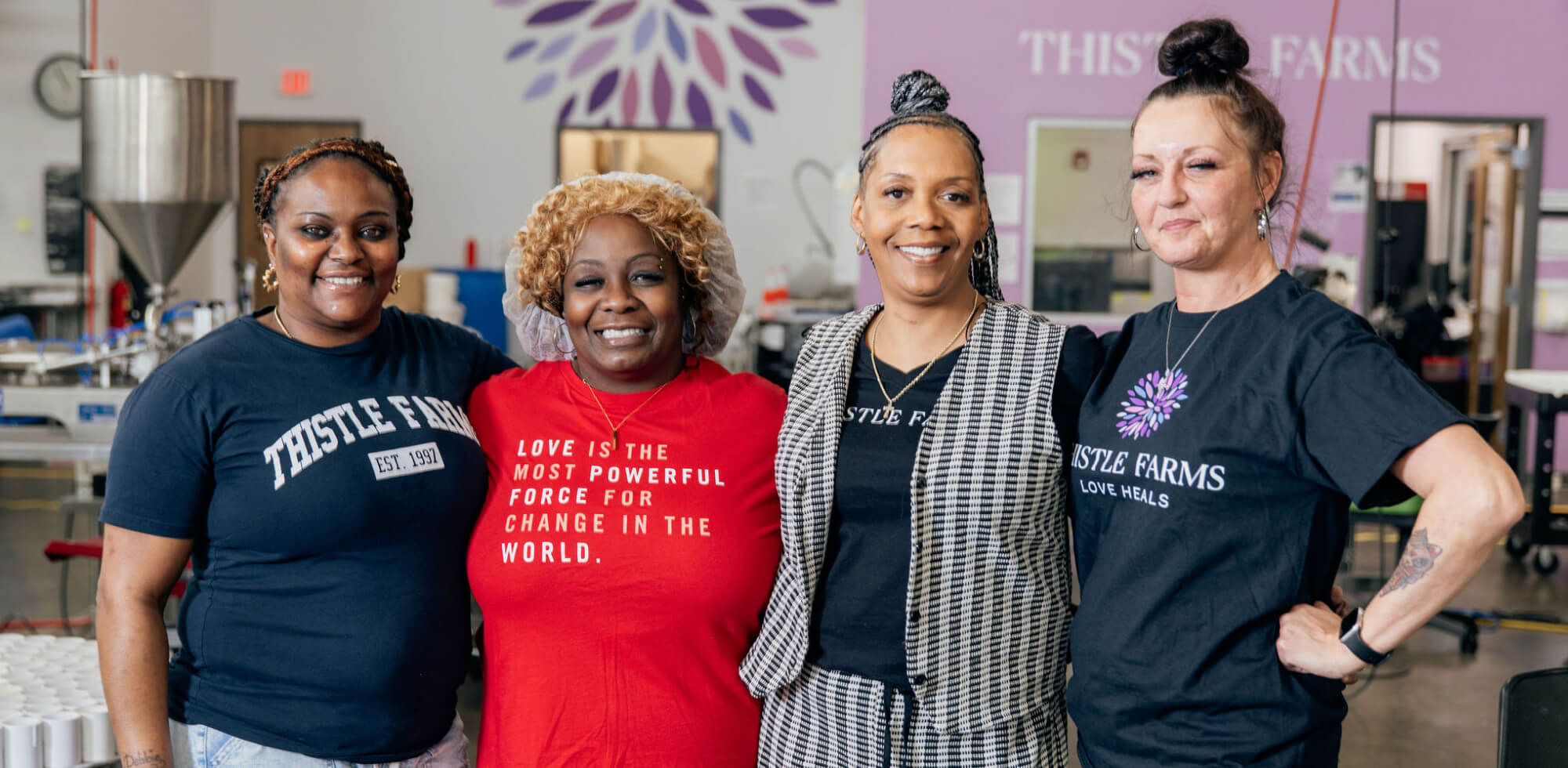 Four women standing together in the Thistle Farms manufacturing facility, wearing 'Thistle Farms' shirts.
