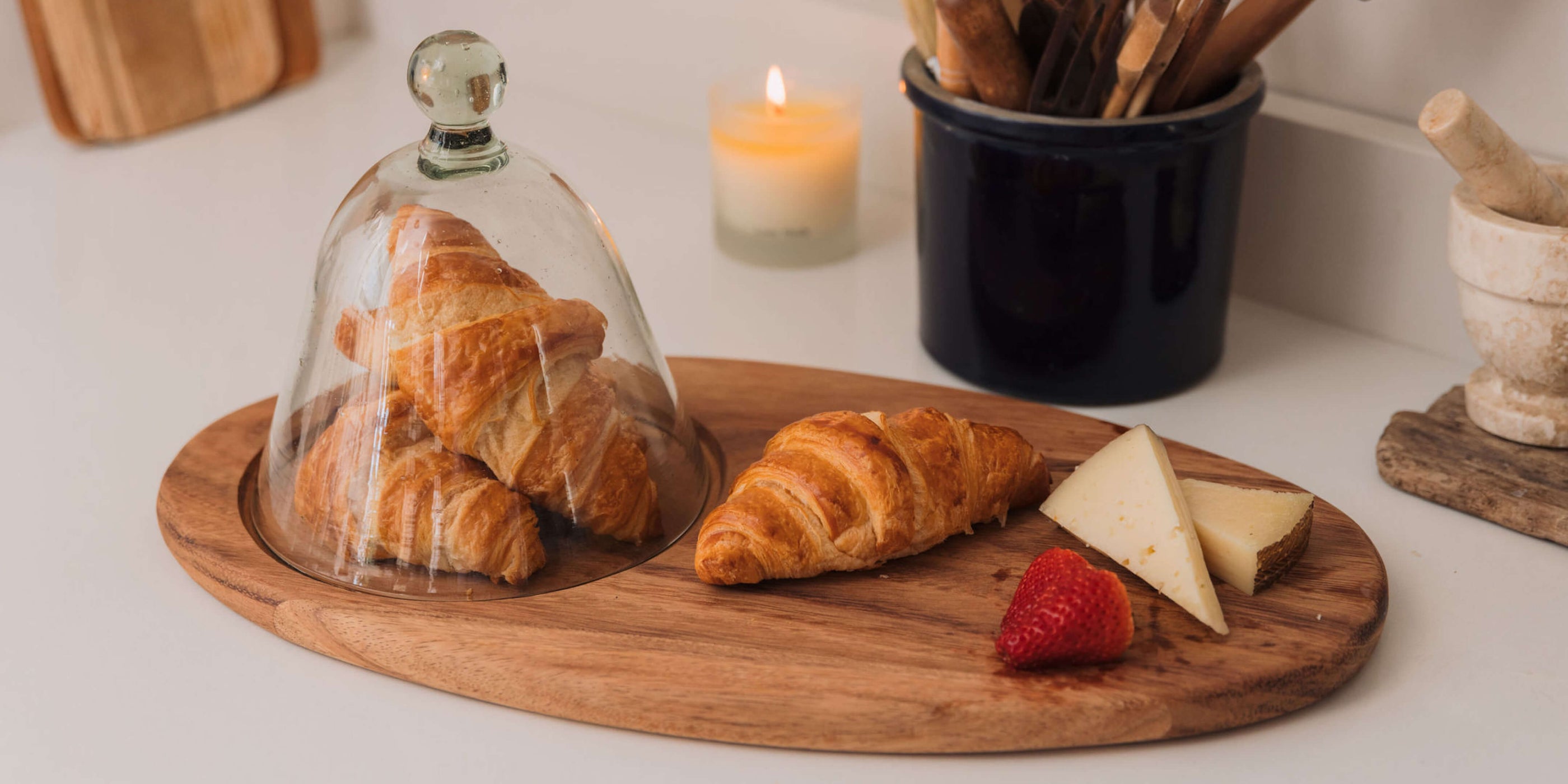 Wooden tray with croissants under a glass dome, cheese, and berries on a white surface.