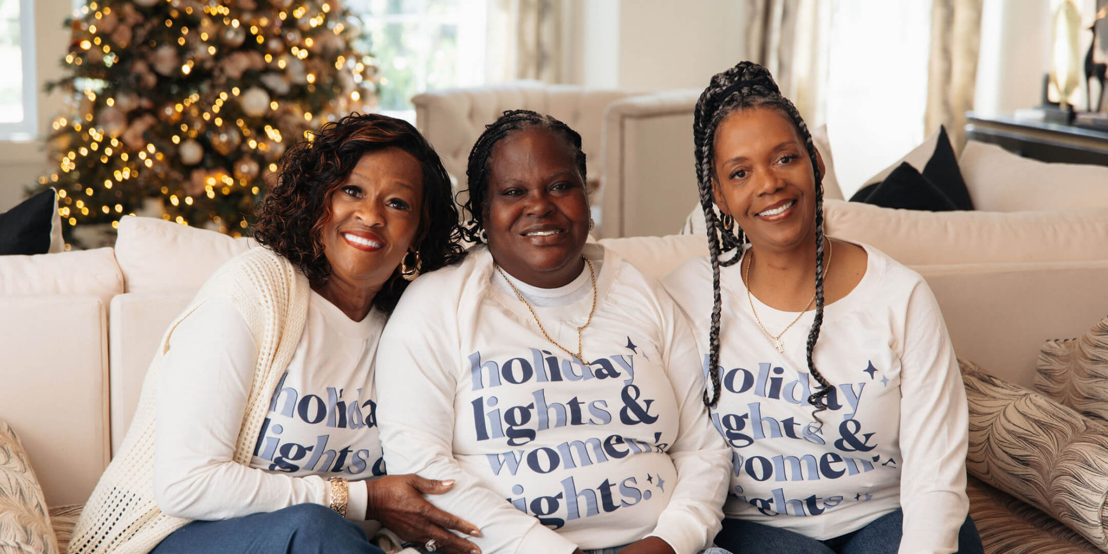 Three women sitting on a couch in front of a decorated Christmas tree.