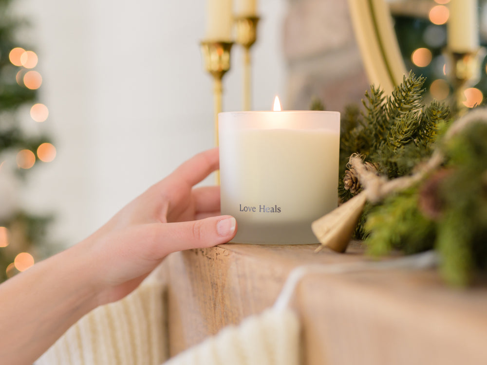 Candle labeled 'Love Heals' held by a hand with festive decorations in the background