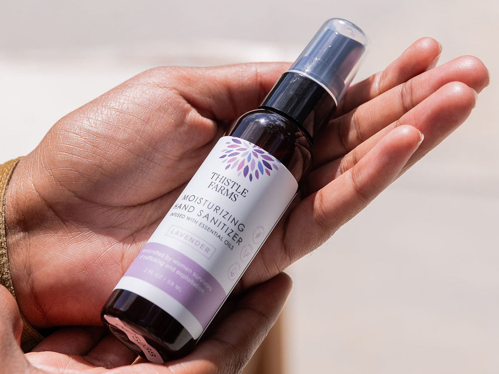 Hand holding a bottle of Thistle Farms lavender hand sanitizer with a white background