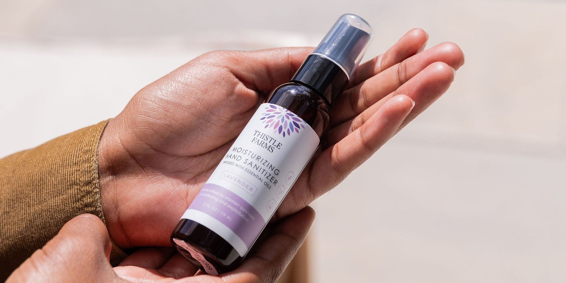 Hand holding a bottle of Thistle Farms lavender hand sanitizer with a white background