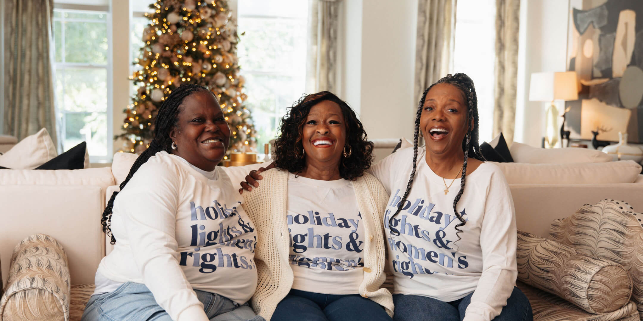 Three women sitting on a couch wearing matching holiday-themed sweatshirts in a cozy living room.