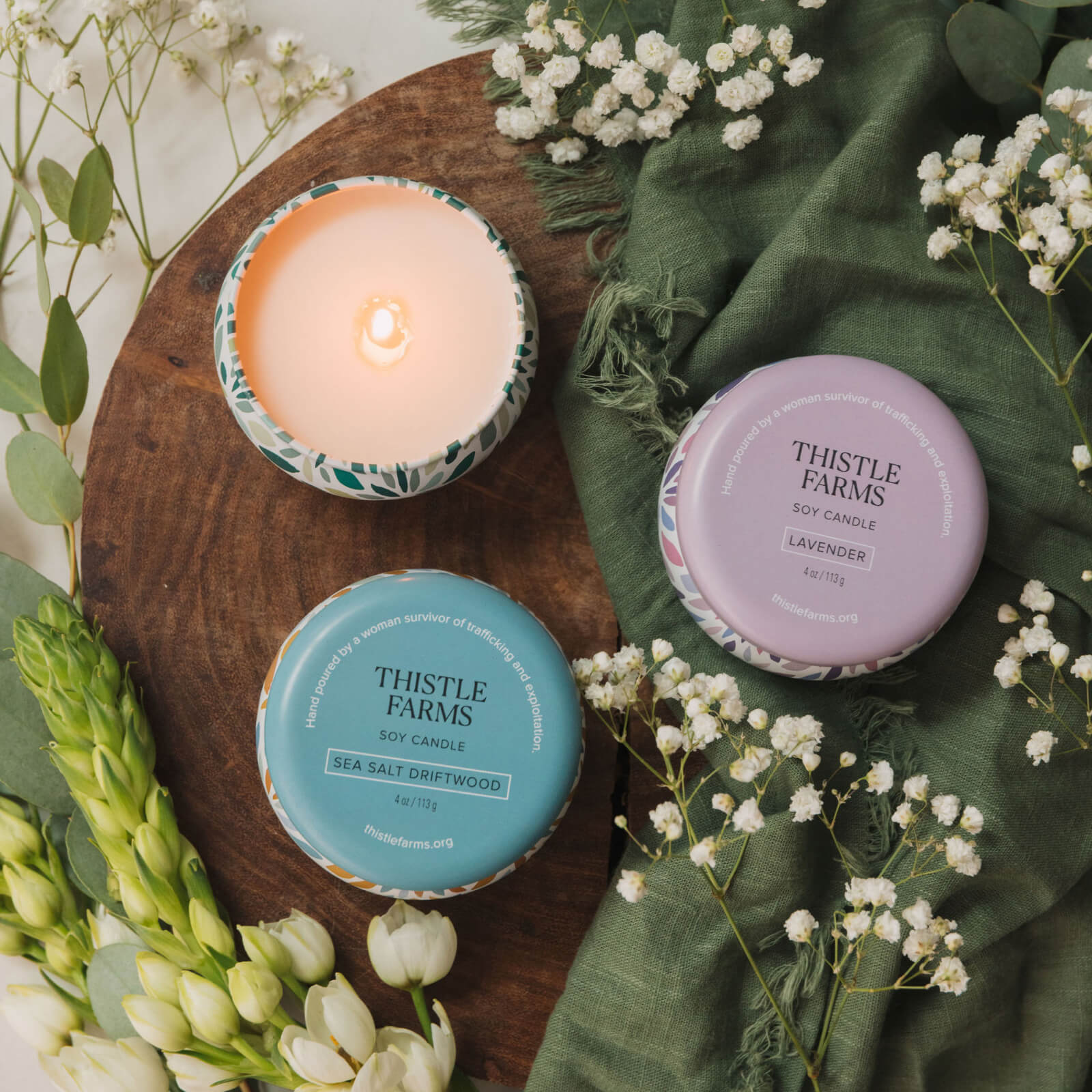 Three Thistle Farms candles on a wooden board with greenery and flowers.