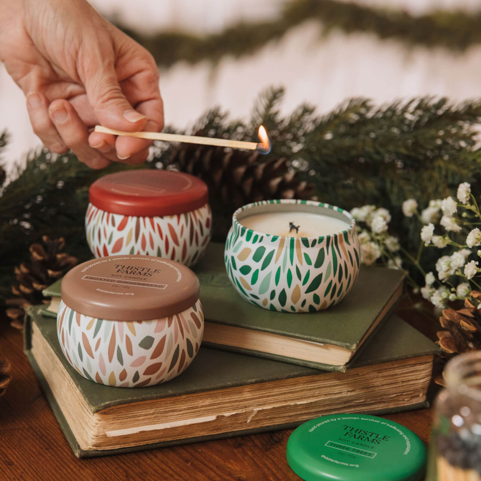 Hand lighting a candle with decorative lids on a stack of books, surrounded by greenery and flowers.