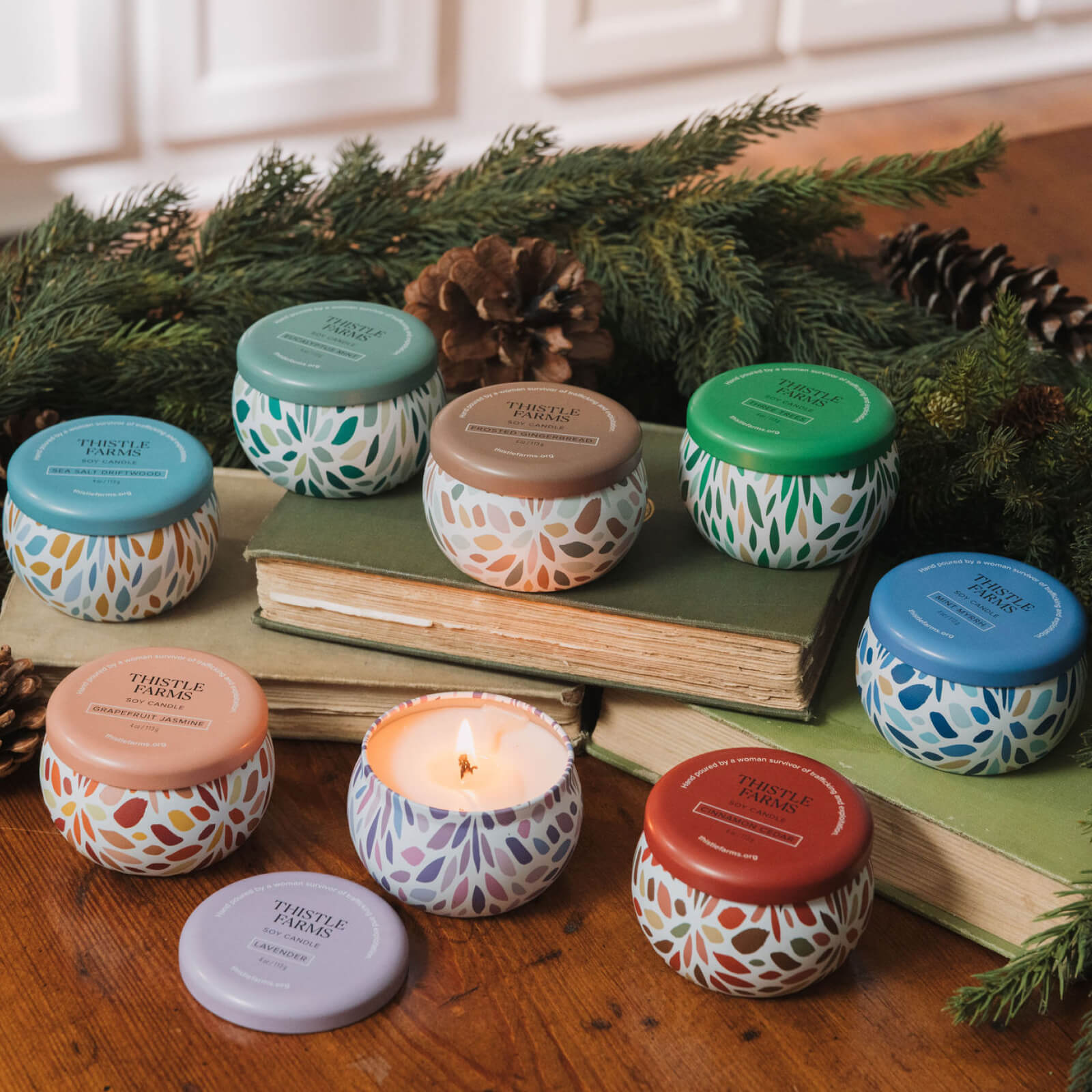 Assorted small candles with colorful lids on a wooden surface with books and greenery.