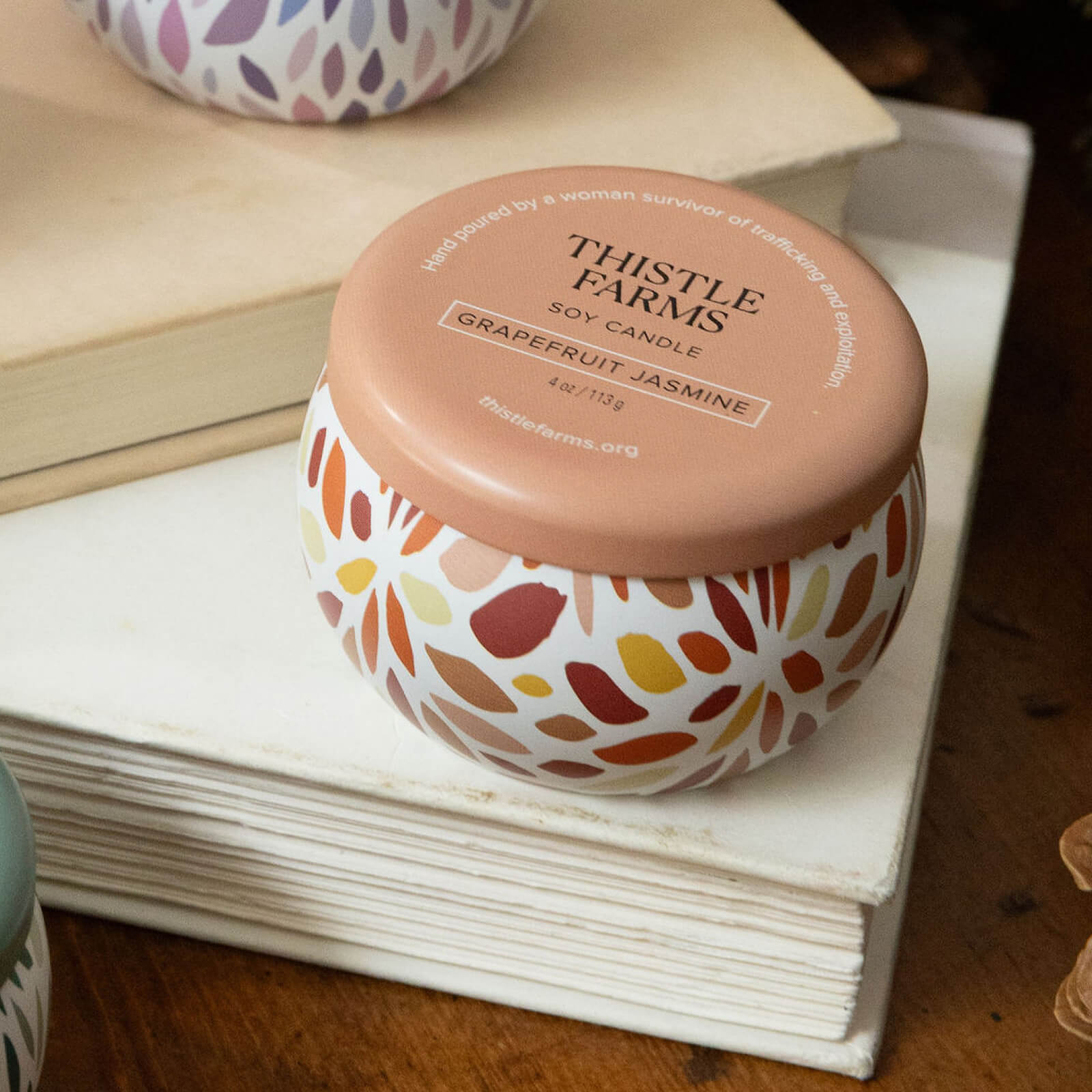 Thistle Farms soy candle with an orange lid and decorative base on a book