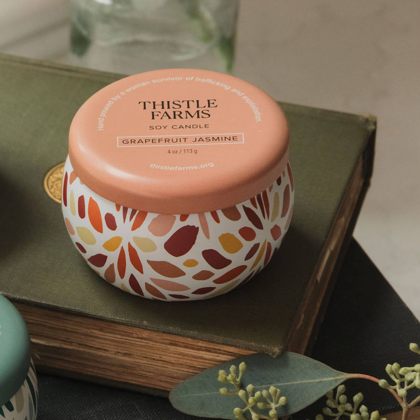 Thistle Farms soy candle with an orange lid and decorative base on a book