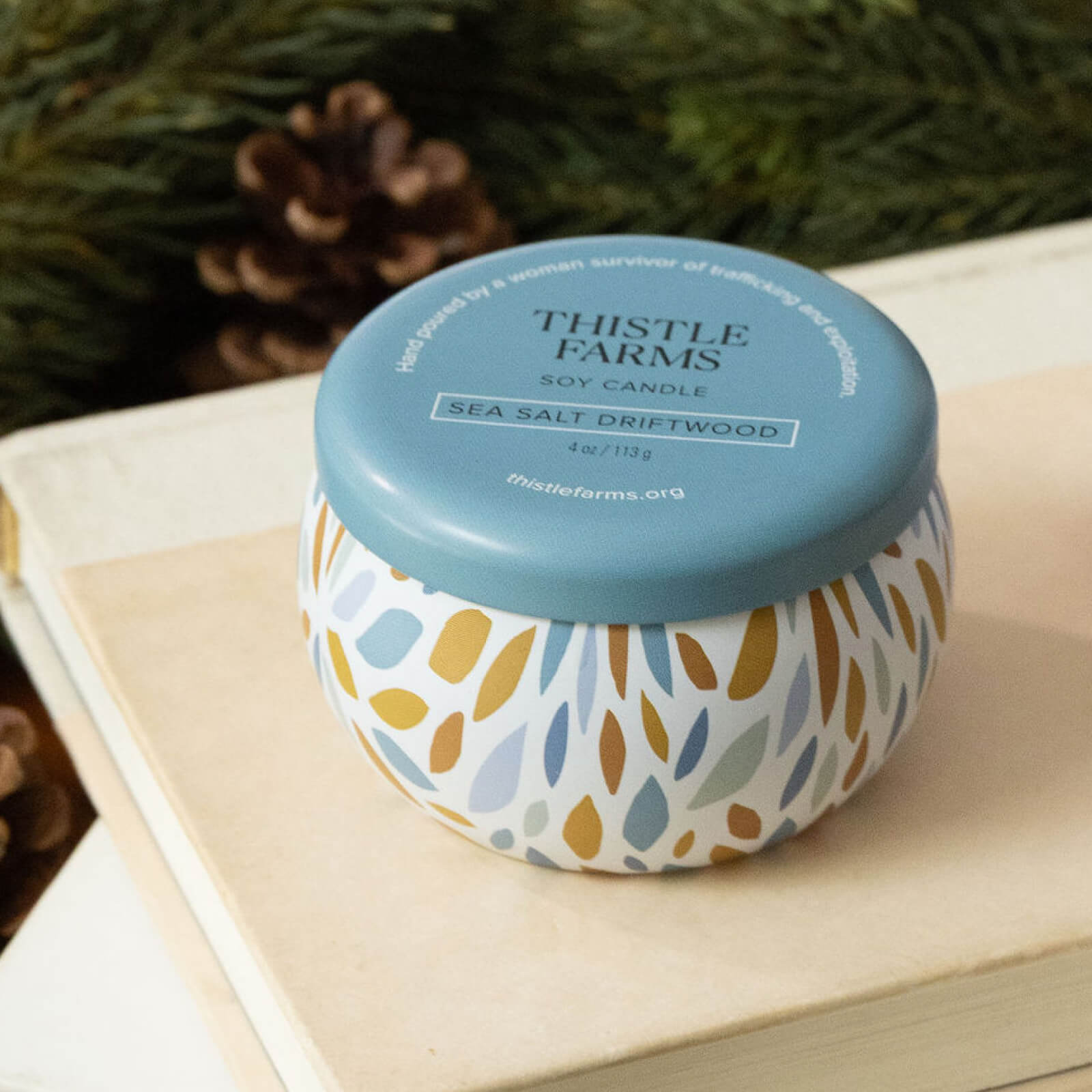 Thistle Farms soy candle with a blue lid and decorative base on a book