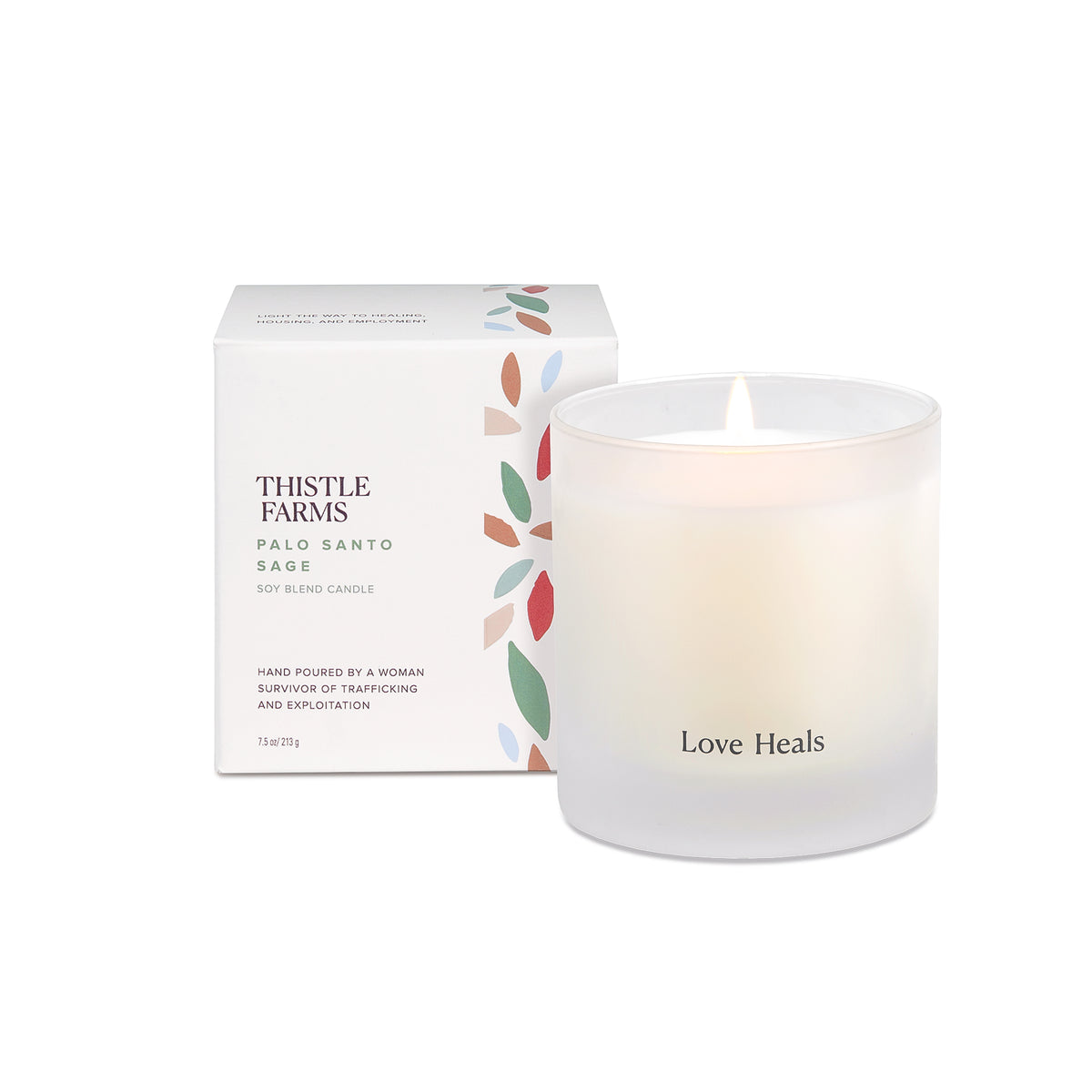 A white candle with a frosted 7.5 oz tumbler packaging labeled 'thistle farms' and 'love heals', with a visible wick and a small flame.