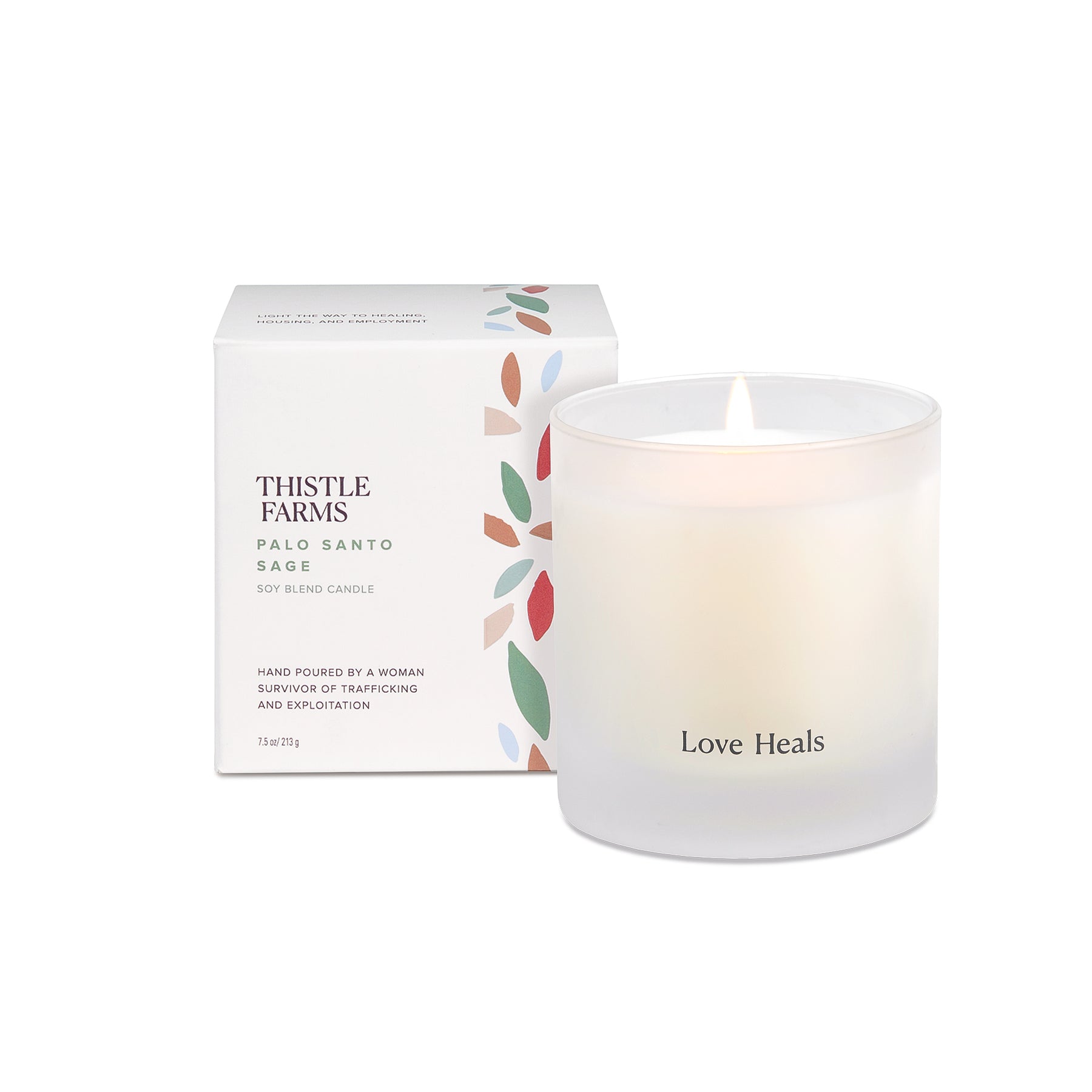 A white candle with a frosted 7.5 oz tumbler packaging labeled 'thistle farms' and 'love heals', with a visible wick and a small flame.