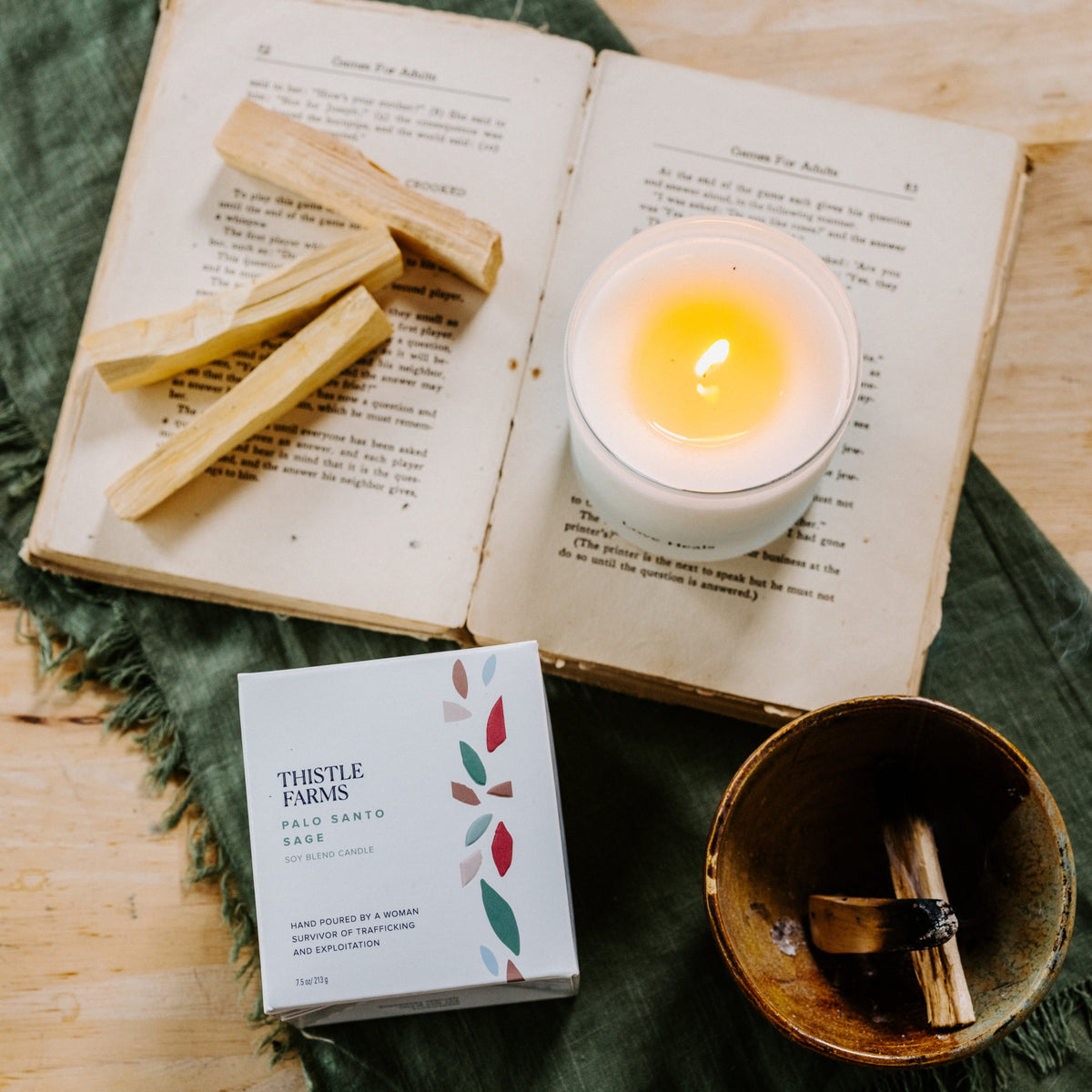 Lit Palo Santo candle placed on a open book with palo santo wood on the other page above a draped green textile on live wood table.