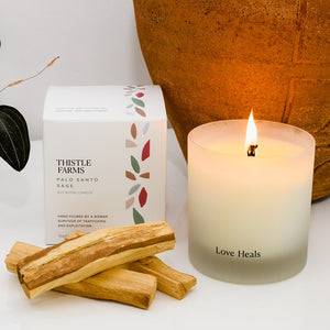 Lit Palo Santo Sage candle next to stacked palo santo wood. The decorated box for the candle touches an orange pot reflecting the glow of the candle.
