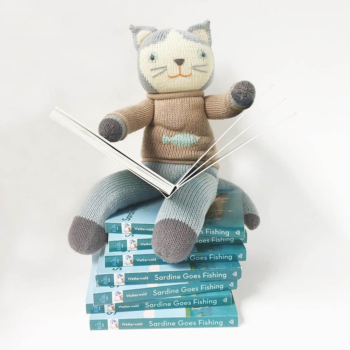 Knitted cat toy sitting on a stack of books titled 'Sardine Goes Fishing' with a white background.