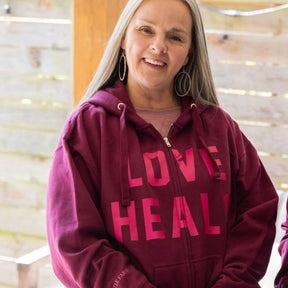 Woman wearing a maroon hoodie with 'LOVE HEALS' text, standing indoors.