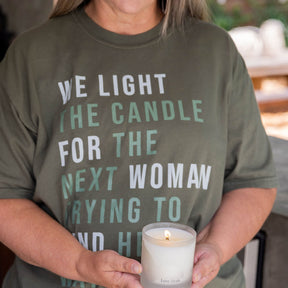Person wearing a green t-shirt with a message "WE LIGHT THE CANDLE FOR THE NEXT WOMAN TRYING TO FIND HER WAY HOME", holding a candle.