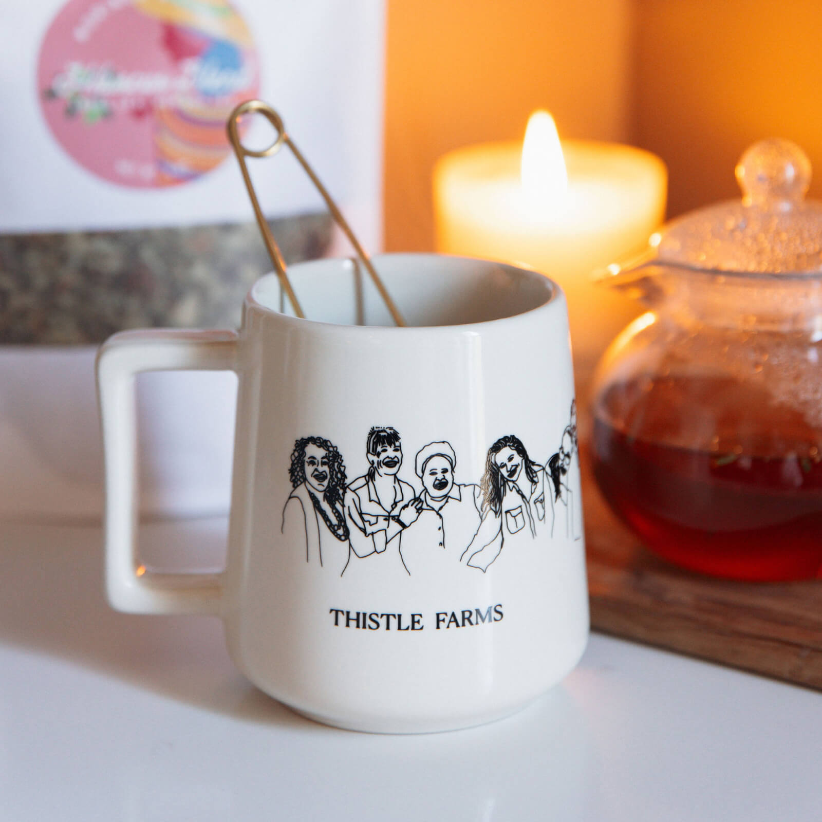 White mug with 'Thistle Farms' design on a table with a candle and teapot in the background