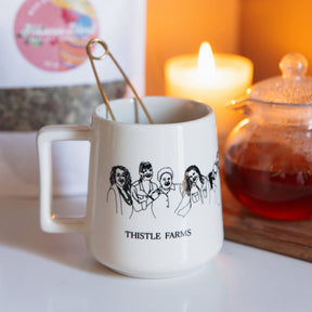 White mug with 'Thistle Farms' design on a table with a candle and teapot in the background