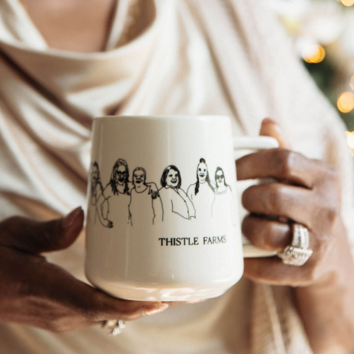 Woman holding Mug that has a design of women embracing with 'Thistle Farms'