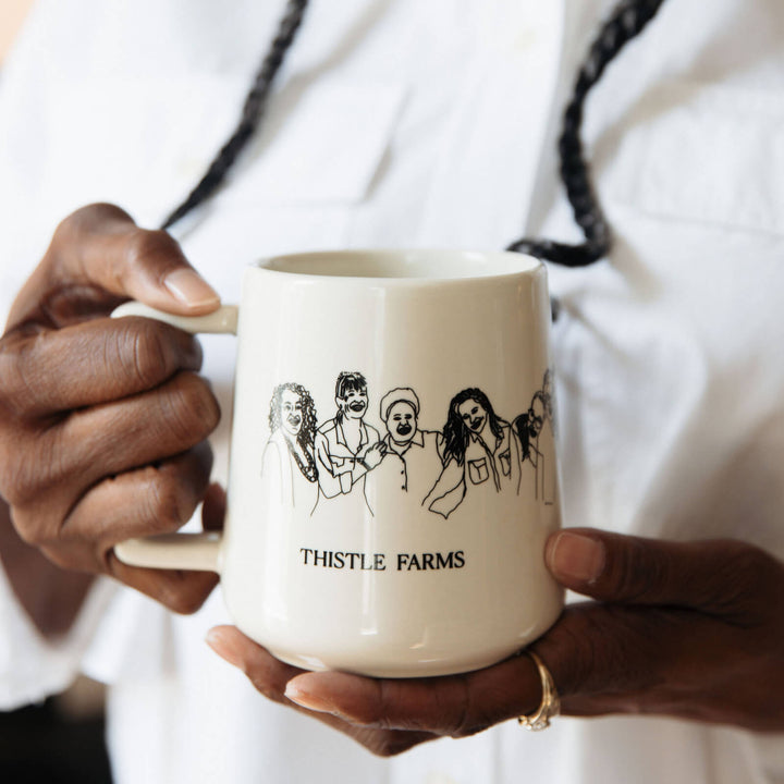 Women is holding Mug has a design of women embracing with 'Thistle Farms'