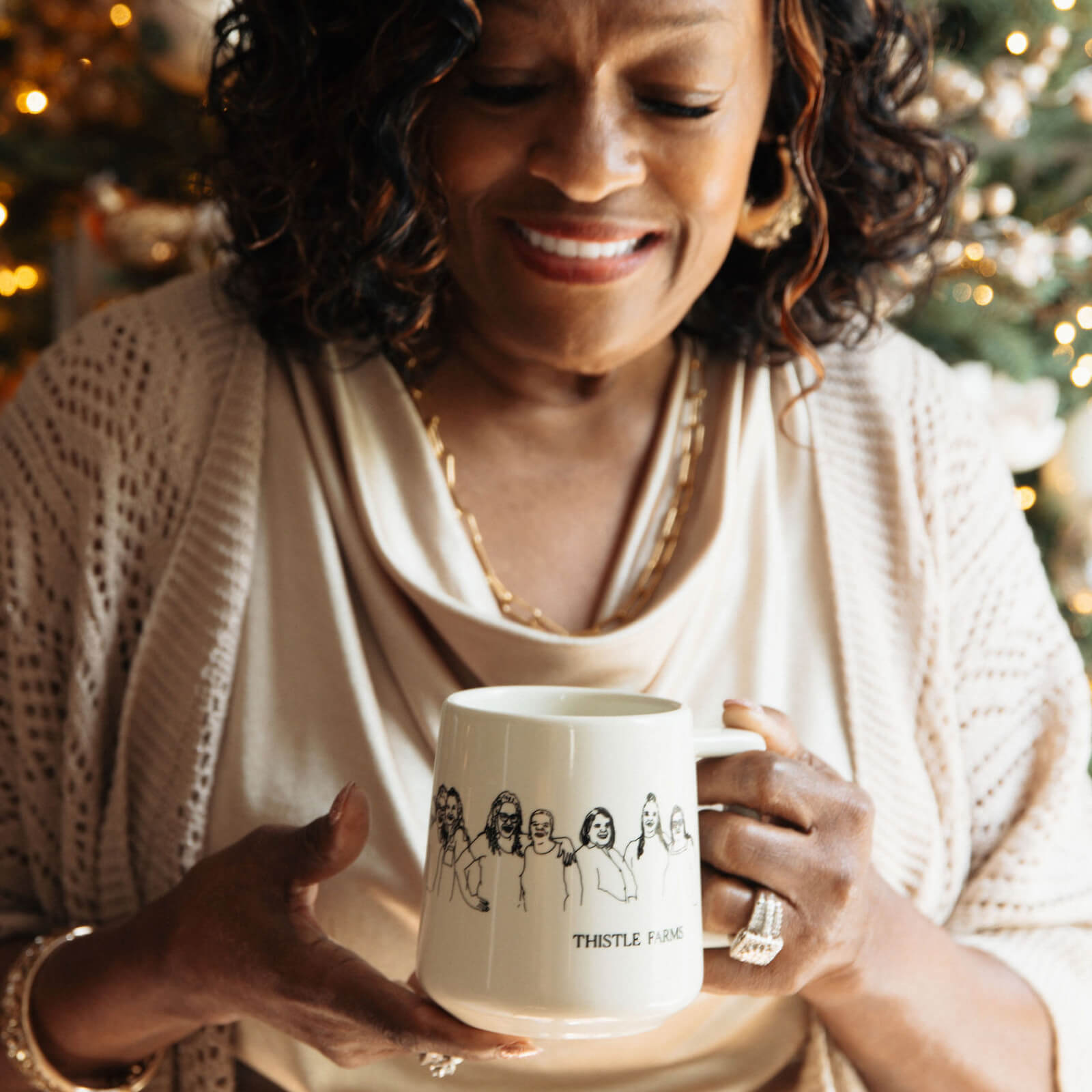 Woman holding Thistle Farms mug with women embracing environment is festive with holiday lights in the background.