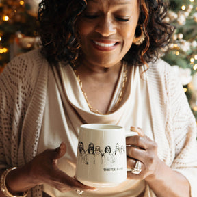 Woman holding Thistle Farms mug with women embracing environment is festive with holiday lights in the background.