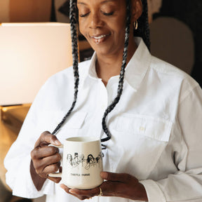 Woman holding a mug with a Thistle Farms mug with women embracing. She is wearing a white shirt in a warm indoor setting.