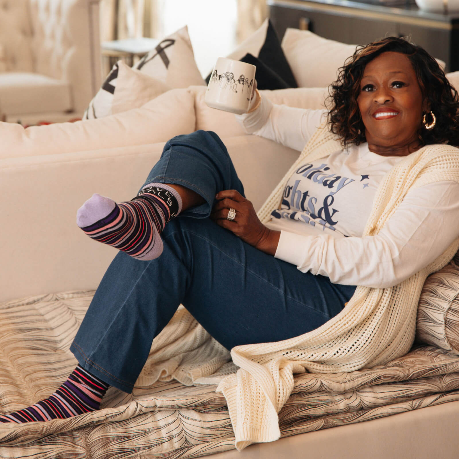 Woman sitting on a couch wearing Love Heals Stripe Socks and a Holiday Lights Womens Rights Tee, with a neutral background.