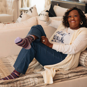 Woman sitting on a couch wearing Love Heals Stripe Socks and a Holiday Lights Womens Rights Tee, with a neutral background.